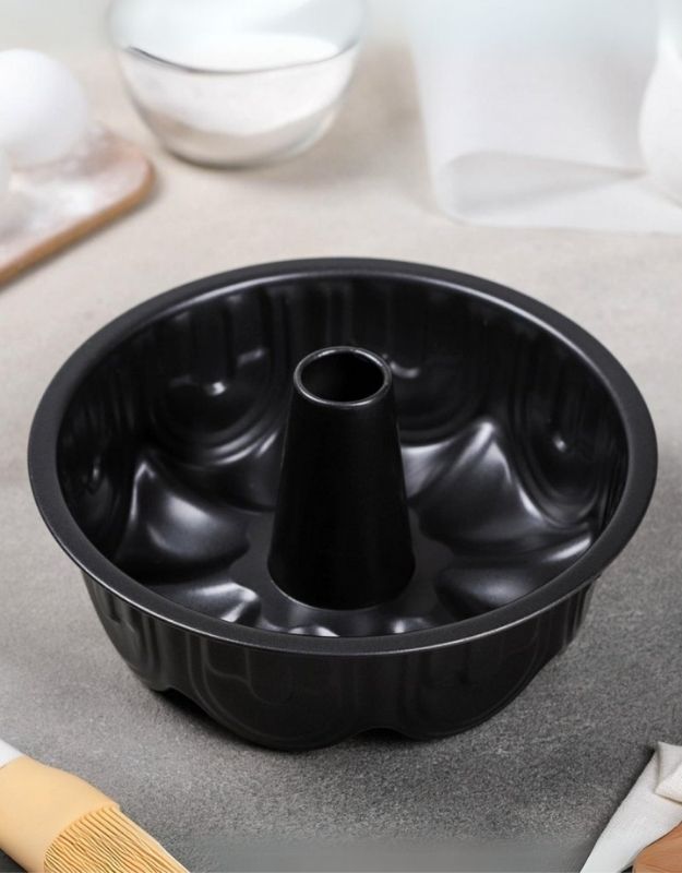 Black Bundt cake pan on a gray surface with kitchen items in the background. Black nonstick bundt cake pan with fluted design, angled view.